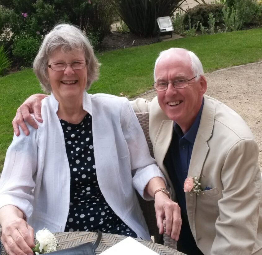 Older man and woman sitting at a table outside, the man has his arm around the woman. Both smiling to camera