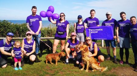 Group of people of all ages in PCUK tops smiling, a lady holding purple balloons and 2 dogs