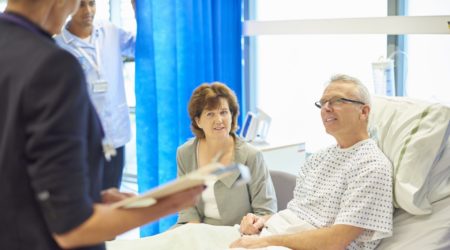 A mature man sits in his hospital bed waiting to listen to a doctor explain his diagnosis whilst being consoled by his wife . In the background a young male nurse adjusts his curtain screen so that he can hear the news in private. They are smiling as the news appears to be good news.