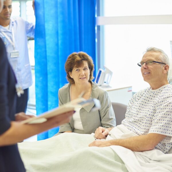 A mature man sits in his hospital bed waiting to listen to a doctor explain his diagnosis whilst being consoled by his wife . In the background a young male nurse adjusts his curtain screen so that he can hear the news in private. They are smiling as the news appears to be good news.