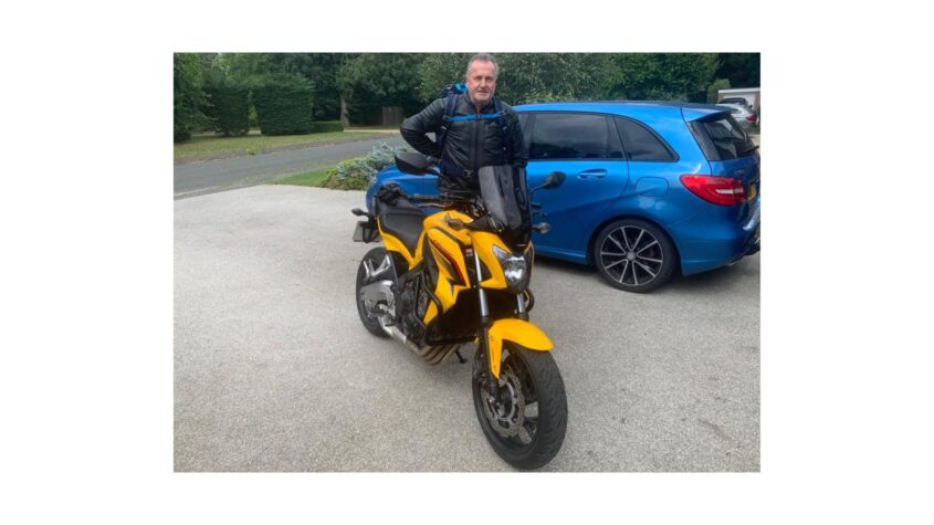Older man standing dressed in biking leathers, standing in front of a bright yellow motorbike