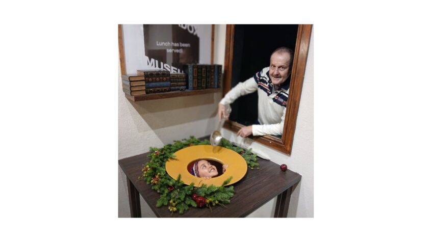 Man pretending to spoon food from a hole in a table, a young girl's face is peeking through the hole
