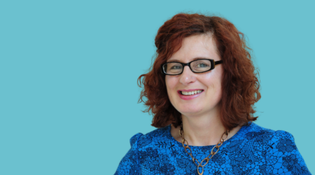 A woman with glasses and curly hair sits in front of a pale blue background