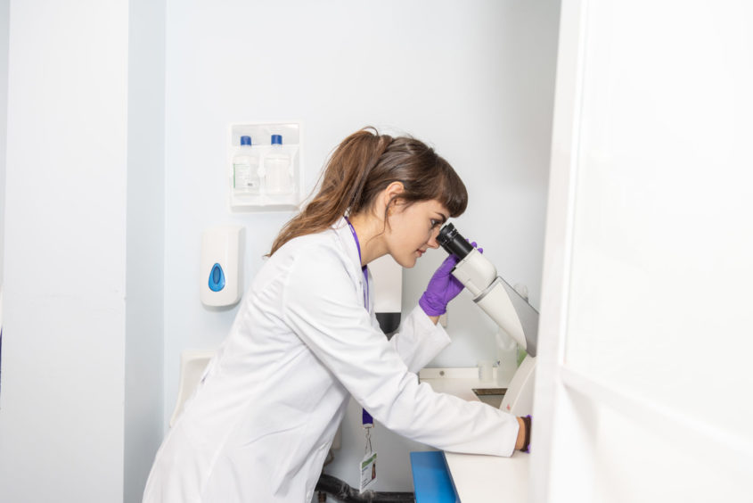 A woman looking through a microscope with purple gloves and a lab coat