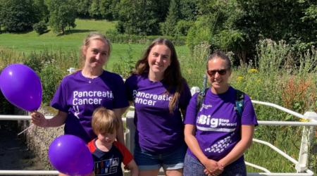 3 women and a child stand in front of a field wearing purple t-shirts and holding purple balloons