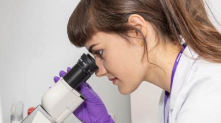 A researcher in a white lab coat looks down a microscope