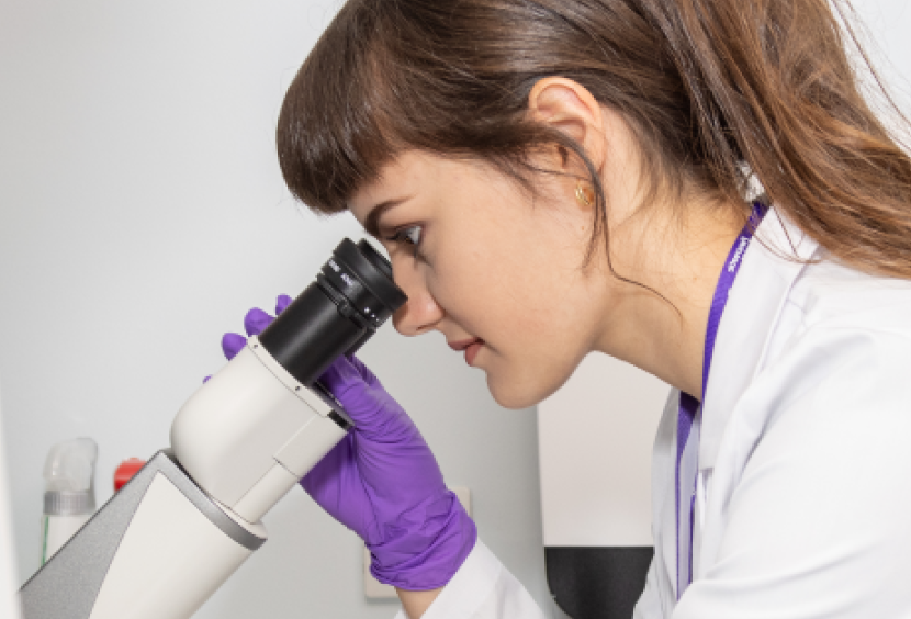 A researcher in a white lab coat looks down a microscope