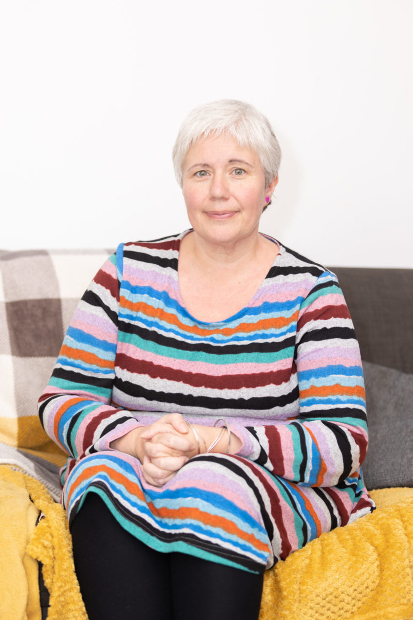 Becki Buggs sits on a sofa in a colourful jumper with her hands clasped