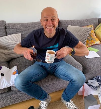 a man sitting on a sofa holding a cup of tea and a muffin with a candle in it - Phil