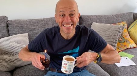 a man sitting on a sofa holding a cup of tea and a muffin with a candle in it - Phil
