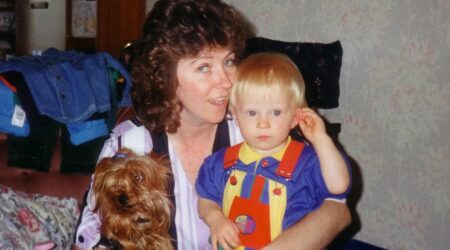 Woman holding small child in one arm and a small dog in the other. Sitting on a sofa looking to camera