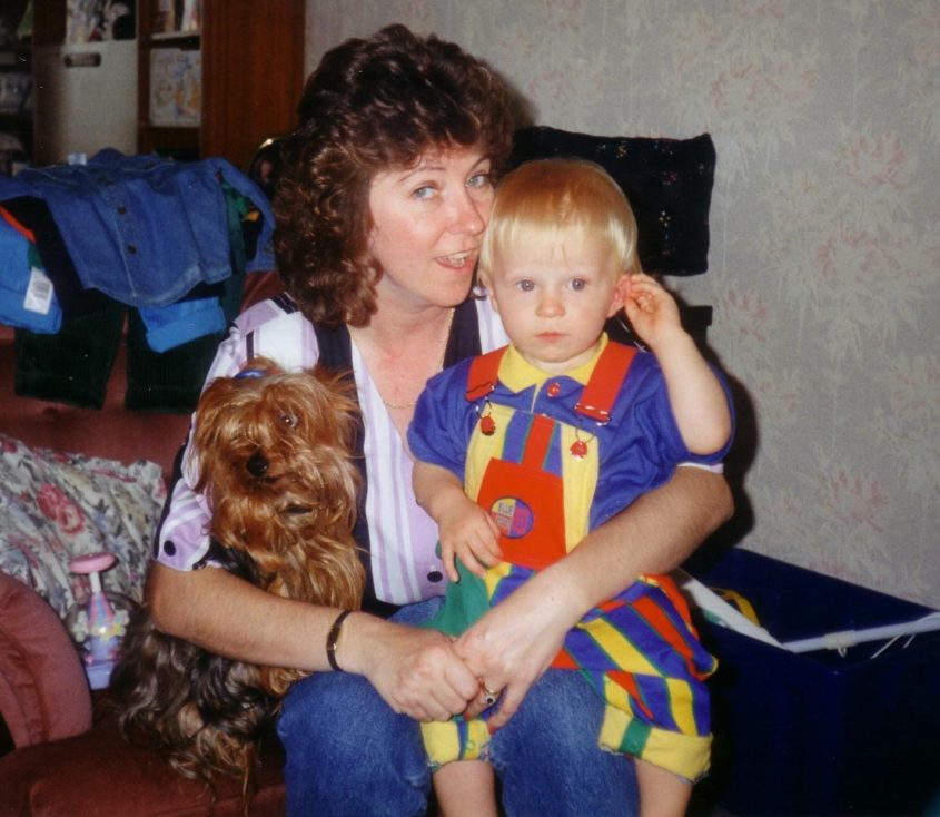 Woman holding small child in one arm and a small dog in the other. Sitting on a sofa looking to camera