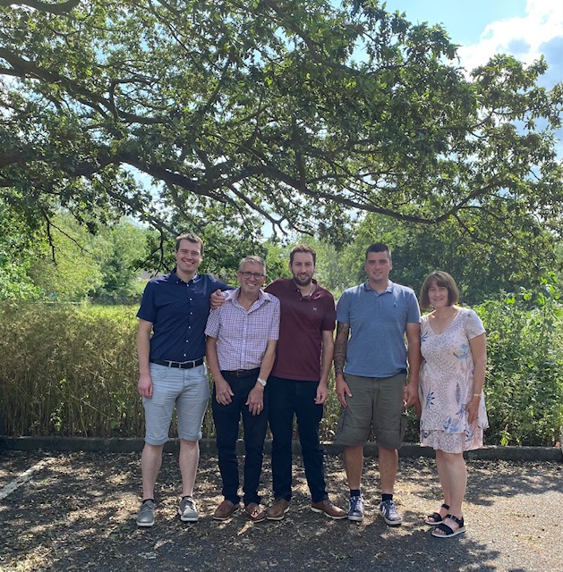 Family of five adults standing in front of some trees