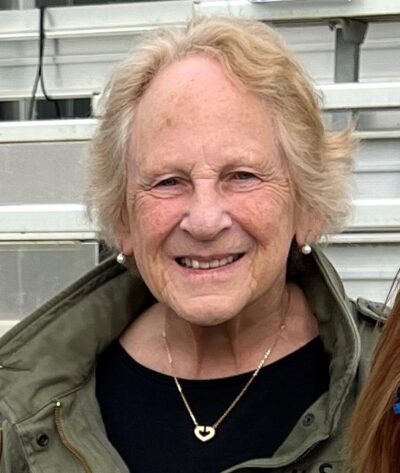 A mature lady wearing a black top with jewellery and khaki jacket smiles at the camera