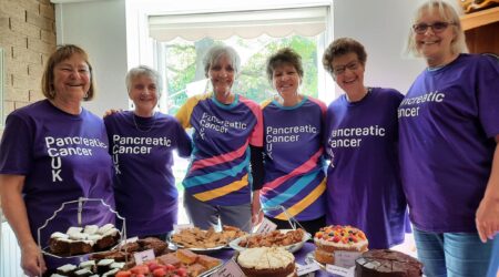 A group of women in purple t-shirts stood around a table, filled with cakes