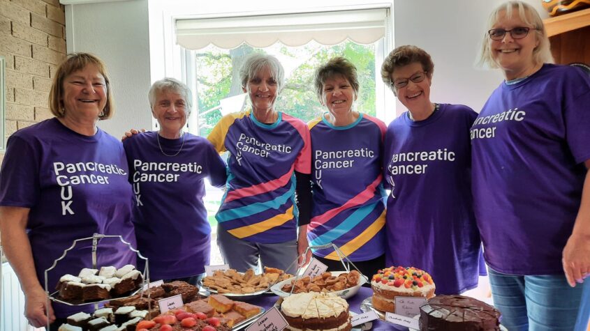 A group of women in purple t-shirts stood around a table, filled with cakes