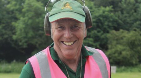 A man in a high-visibility vest and ear defenders smiles on a clay pigeon shoot