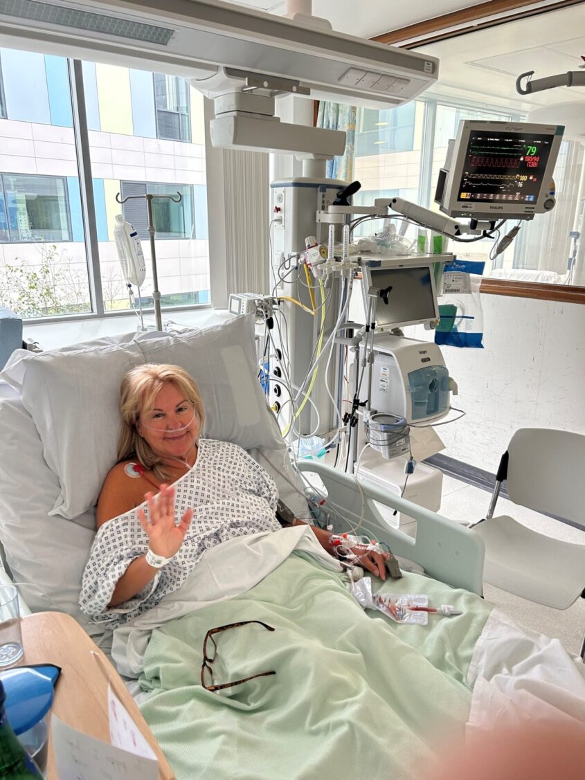 Pippa in a hospital bed, connected to various machines smiling and waving to camera