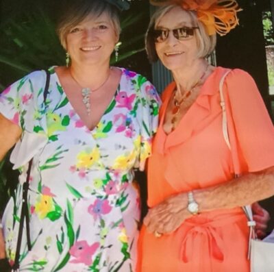 Mother and daughter standing with arms around each other wearing brightly coloured dresses for a summer wedding
