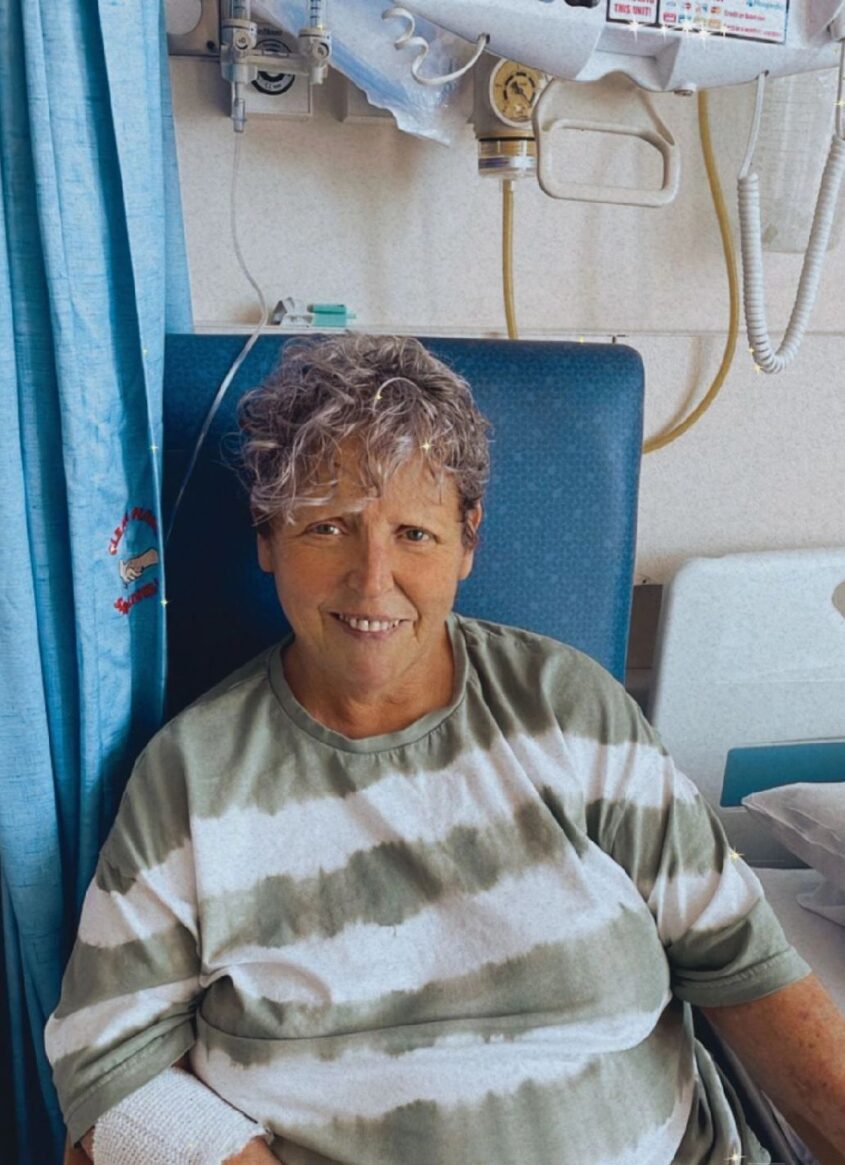 Woman sitting in a chair in a hospital ward, wearing striped top and smiling to camera