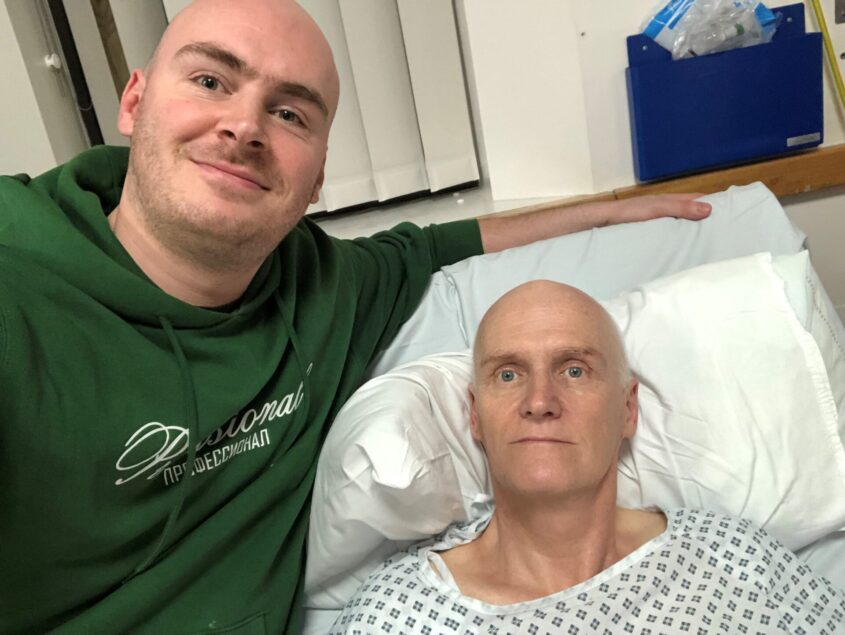 Lewis stands with his arm around a hospital bed, where his dad, Jeff is lying.