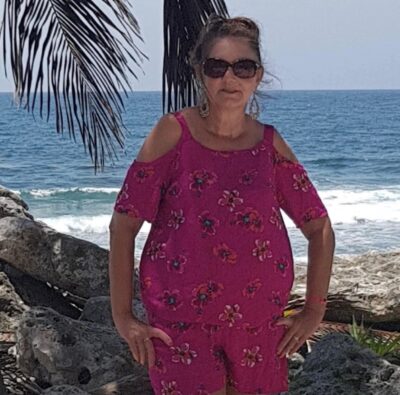 woman in sunglasses standing in front of the sea, underneath a palm tree