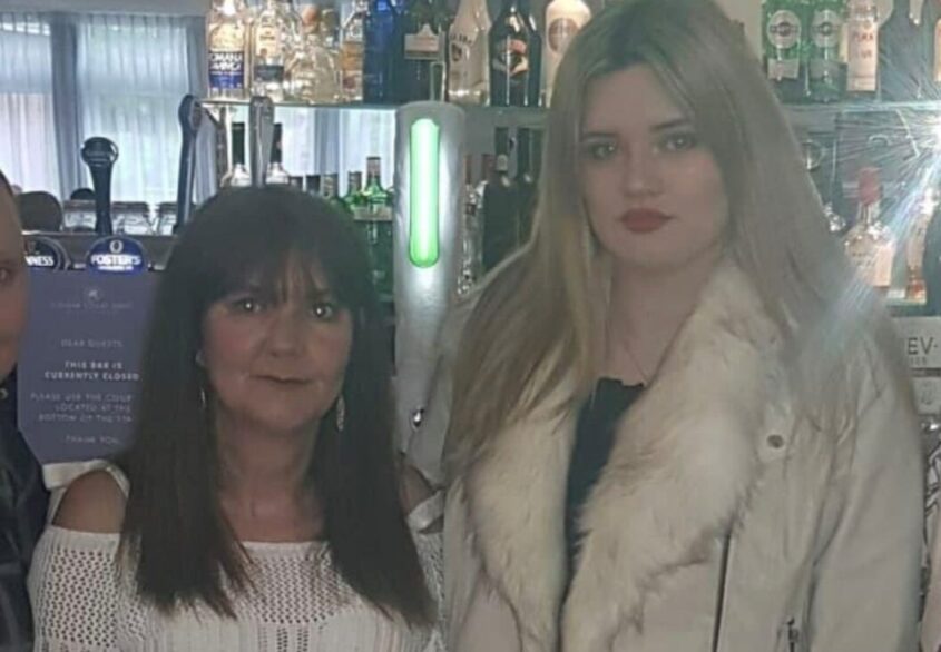 One older and one younger woman standing in a bar, looking to camera