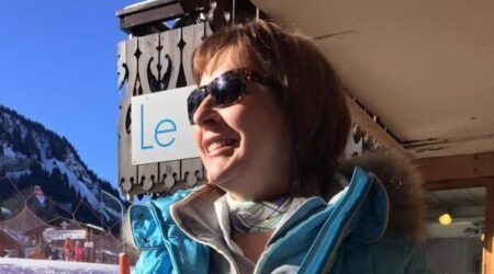 Woman standing in front of ski slopes wearing ski gear and shades and smiling