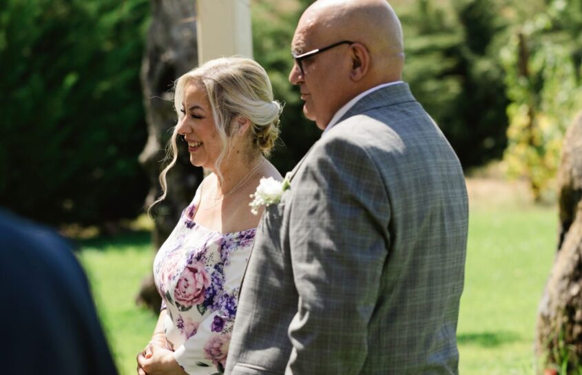 Older man and woman dressed smartly for a wedding. .Standing outside and looking away from camera, smiling.