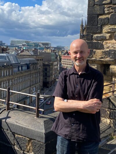 Volunteer Ollie standing in front of some buildings, smiling at the camera