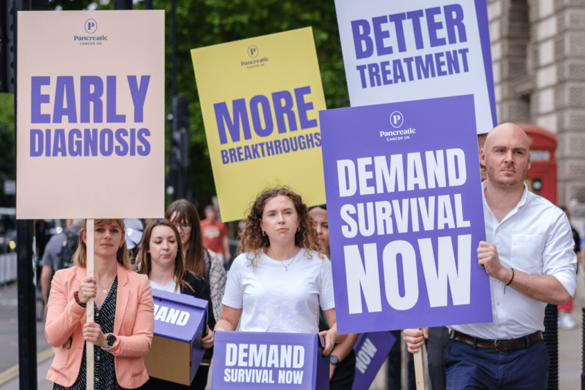 Team of PCUK staff with signs saying 'early diagnosis', 'more breakthroughs', demand survival now' and 'better treatment', walking towards Downing Street.
