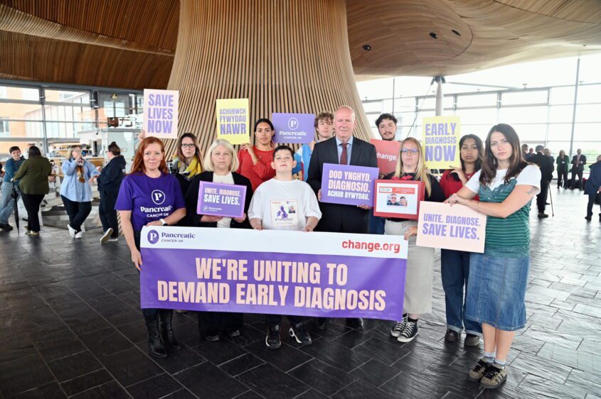 A group of people indoors at the Senedd hold signs advocating for early diagnosis of pancreatic cancer. The mood is urgent and united.