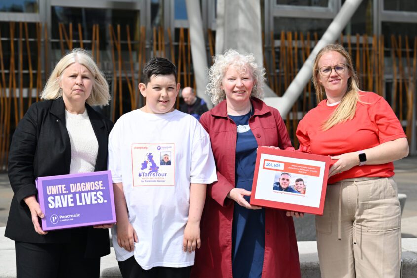 A group of four, two of whom are holding boxes filled with signatures, stand outside Scottish parliament. They are calling for early detection of pancreatic cancer