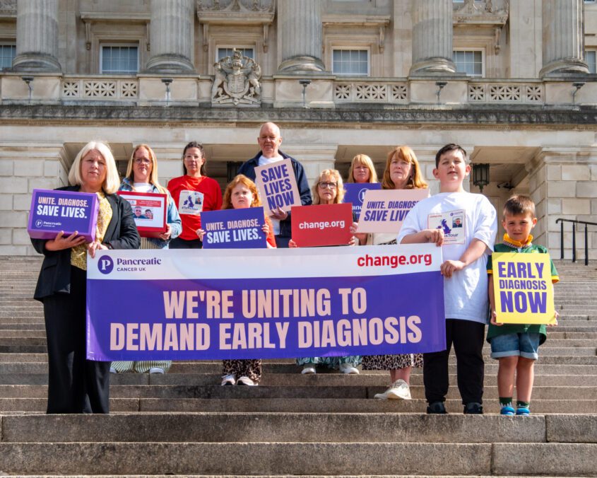 Group of ten stand on stairs outside Stormont, holding signs advocating for early detection of pancreatic cancer to save lives