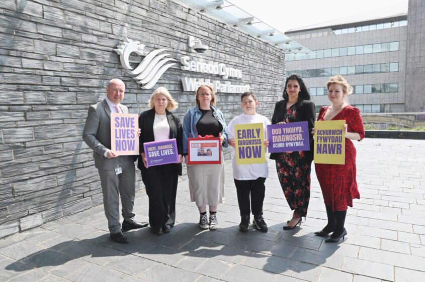Group of six people stand outside the Welsh Parliament, holding signs advocating for early diagnosis of pancreatic cancer to save lives