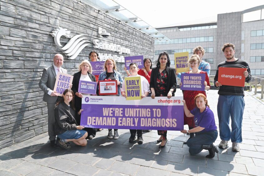Group of twelve people stand outside the Welsh Parliament, holding signs advocating for early diagnosis of pancreatic cancer to save lives