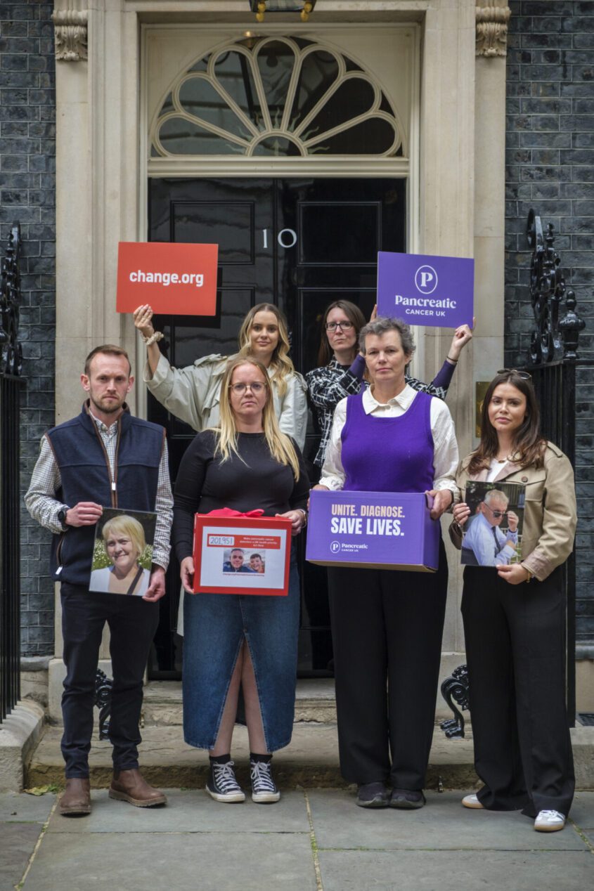 Group of six people stand outside Number 10, Downing Street. Two people at the back hold signs calling for early detection of pancreatic cancer. Two people at the front hold boxes that include signatures from people calling for early detection.