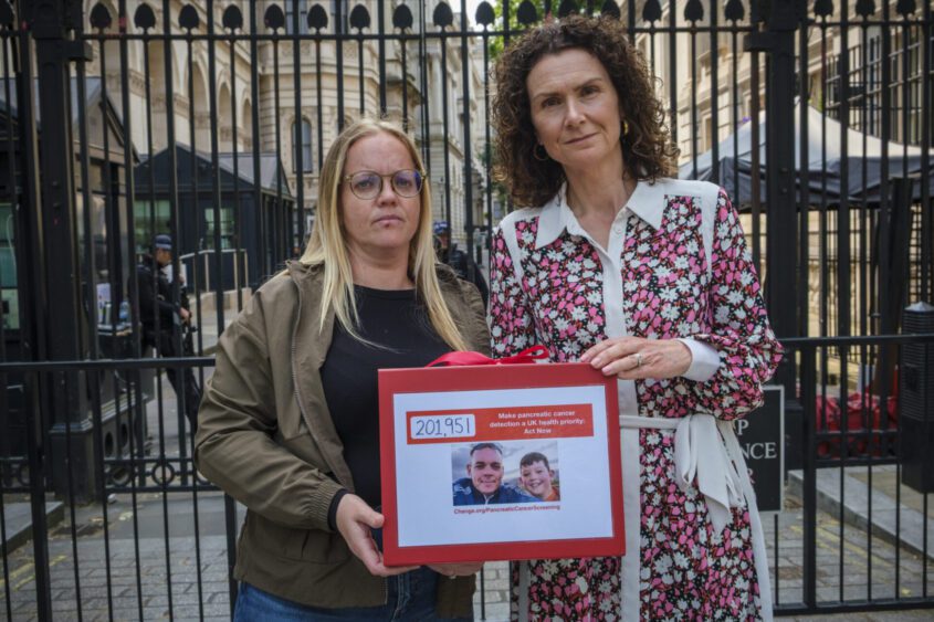 Two women stand in front of a gate, holding a petition box including over 200,000 signatures calling for early detection of pancreatic cancer