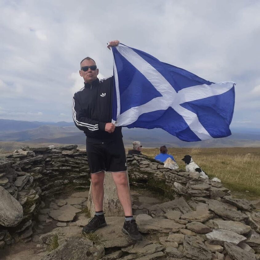 Man stands on rocky landscape, holding the flag of Scotland proudly,
