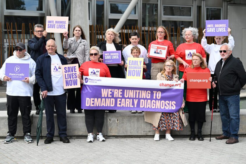 A group of fourteen, including two children, stand outside Scottish parliament, holding signs calling for early detection of pancreatic cancer to save lives.