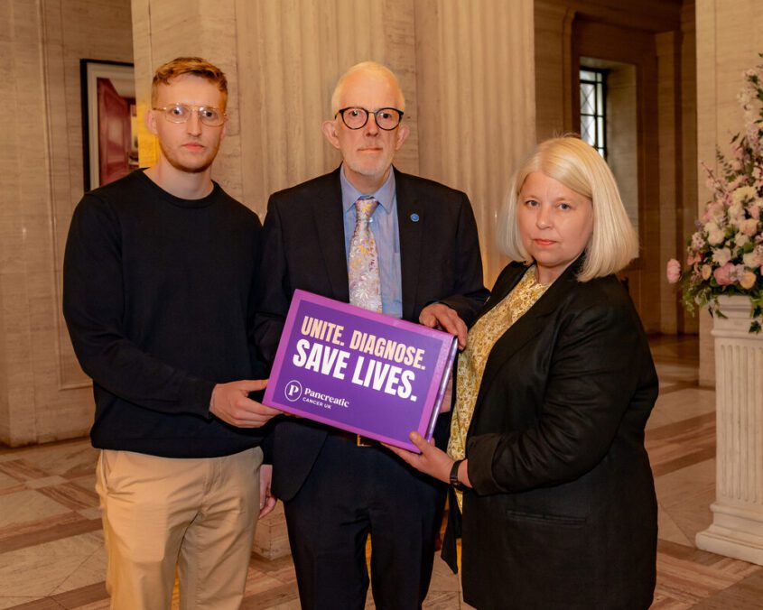 Three individuals stand together in a formal setting, holding a purple sign that reads