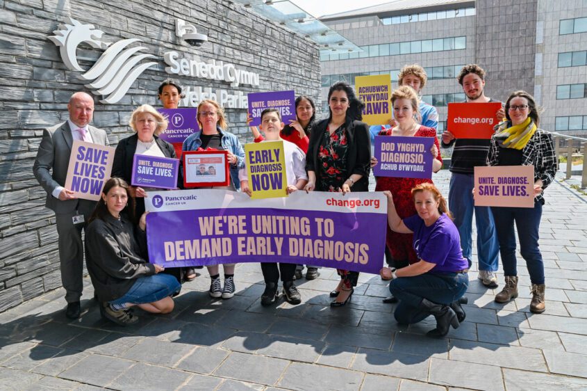 A group of people holding signs advocating for early diagnosis and saving lives, gathered outside a healthcare facility, promoting awareness and support for their cause.