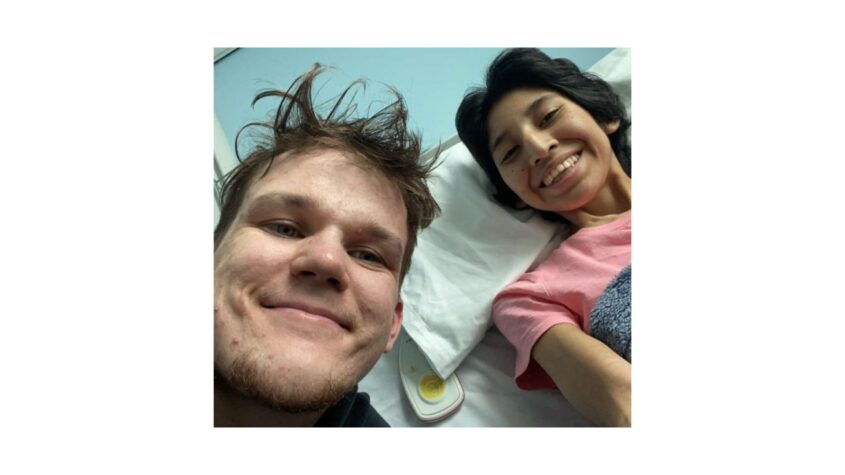 Selfie of young woman lying in hospital bed with young man beside her