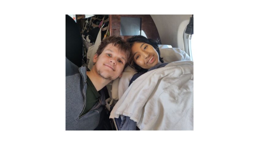 Young woman lying on medical bed on a plane with young man sitting beside her
