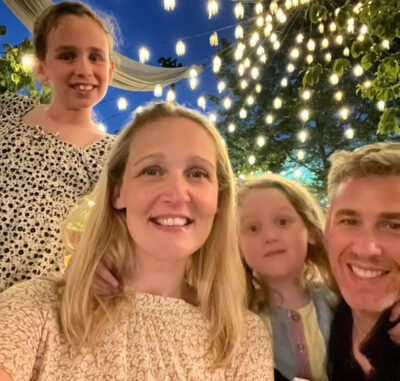 Mum, Dad and 2 young girls standing in front of fairy lights