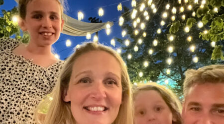 Mum, Dad and 2 young girls standing in front of fairy lights