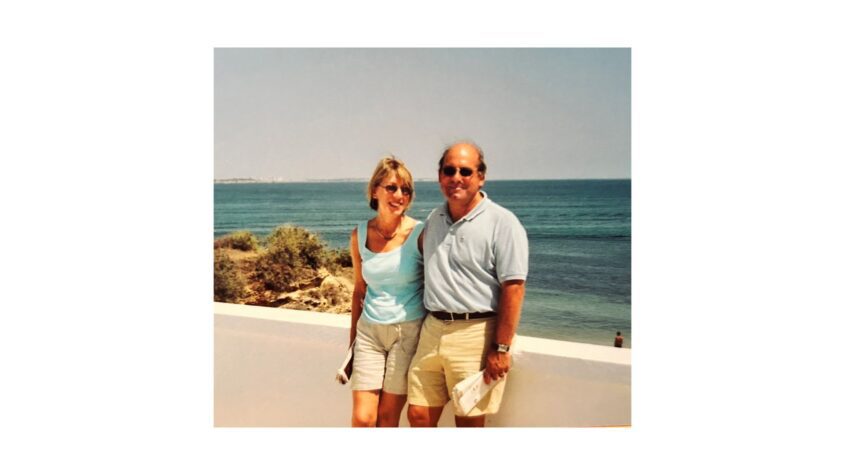 Couple wearing summer clothes, standing in front of a low white wall with sea in the background