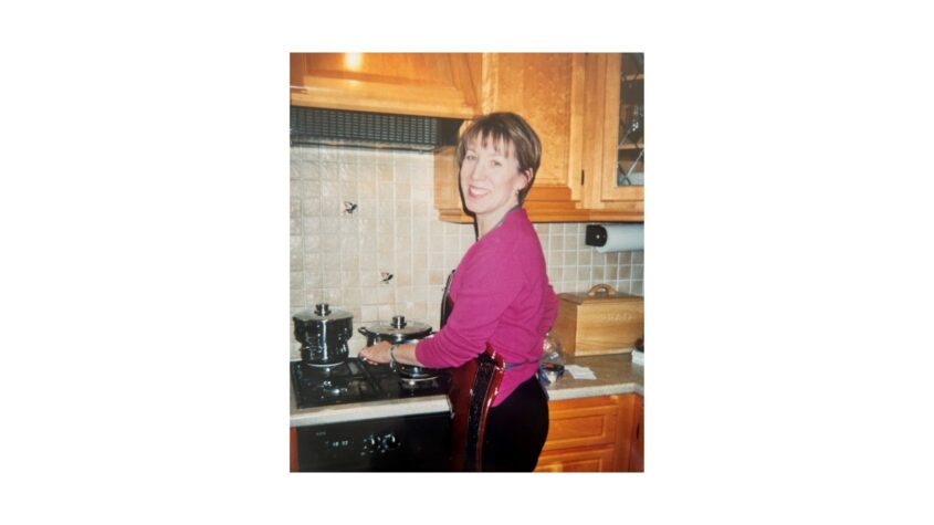 Woman wearing pink blouse standing in front of a cooker in the kitchen looking back over her shoulder to camera