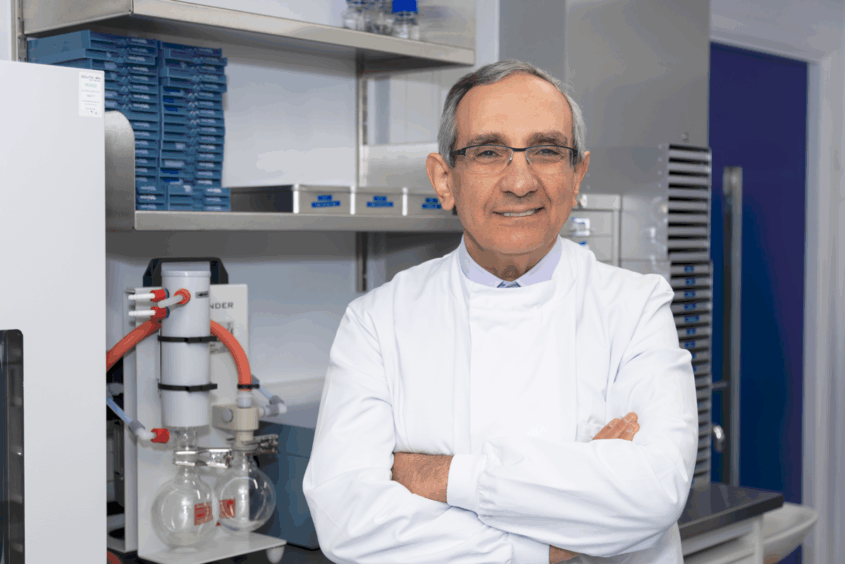 A smiling scientist in a lab coat stands confidently with arms crossed, surrounded by laboratory equipment and shelves filled with blue storage containers.