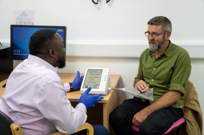 A man wearing gloves demonstrating a breath collection device to another man sat at a desk